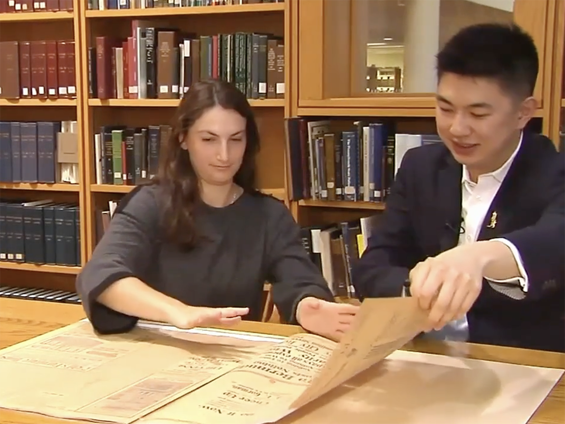 Two college students sit in a library and handle a fragile archival newspaper carefully